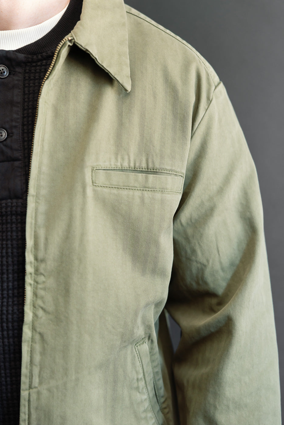 Close-up side view of an olive green canvas jacket showing the chest pocket and fabric texture.