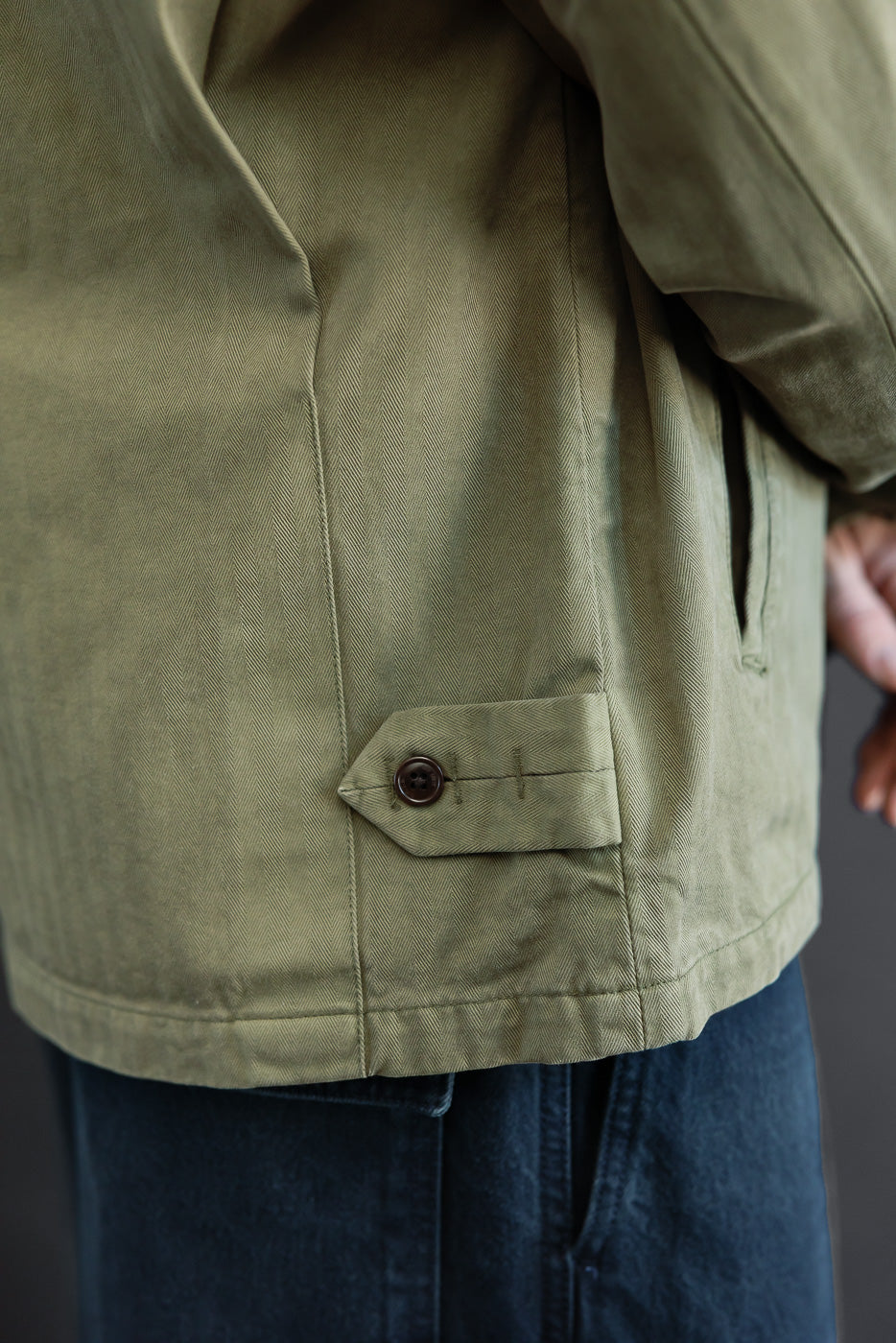Close-up of the back hem and side tab detail on an olive green canvas jacket worn by a male model.