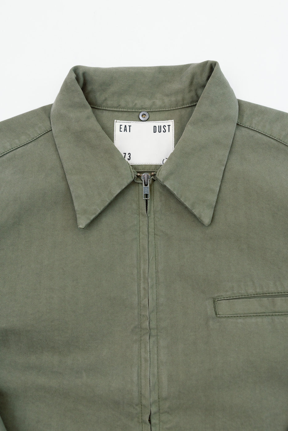 Close-up front view of olive green jacket showing collar, zipper, and Eat Dust neck tag.