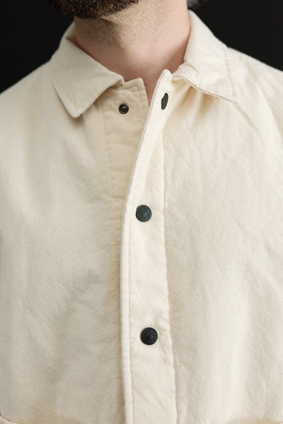 Close-up front view of ecru jacket on male model, showing collar, snap buttons, fabric texture.