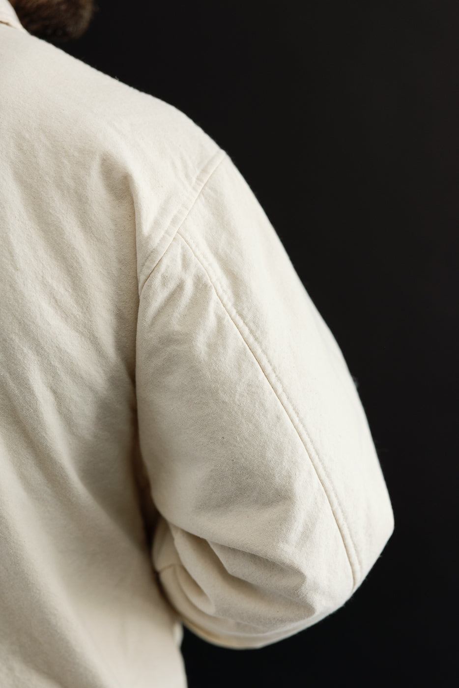 Close-up of male model's right arm and back shoulder in ecru canvas jacket, showing fabric texture and seam details; model is 5'9