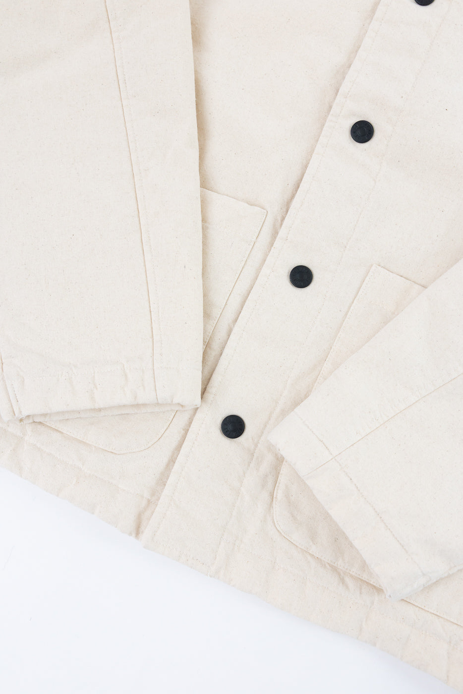 Close-up of ecru canvas jacket front, showing sleeve, matte black buttons, and hip pockets.