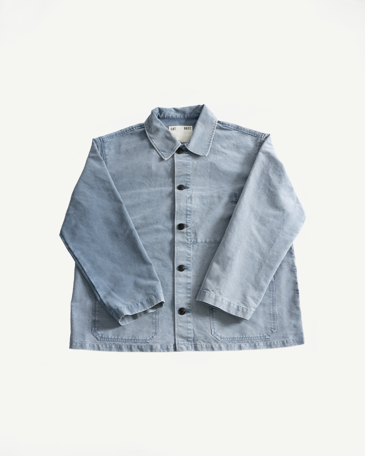 Light-wash denim chore jacket, titled M-B-006 - Coach Jacket Nebraska Cotton - Coastal Blue, featuring a pointed collar, front button-up closure, patch pockets, and “EAT DUST” label, crafted for relaxed everyday wear.