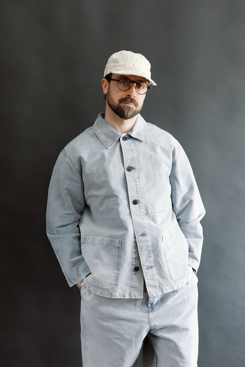Man wearing M-B-006 Coach Jacket Nebraska Cotton in Coastal Blue, featuring a relaxed fit, four pockets, and slightly dropped shoulders, styled with matching trousers and a lightly textured off-white cap.