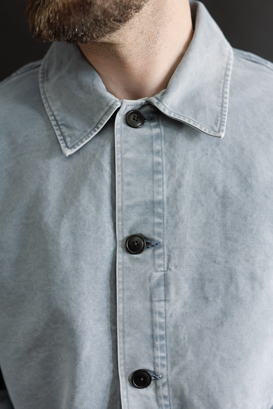 Close-up of M-B-006 - Coach Jacket Nebraska Cotton - Coastal Blue, showcasing its relaxed fit, unique texture, and black buttons against a stubbly beard backdrop, embodying Dant's premium menswear style.