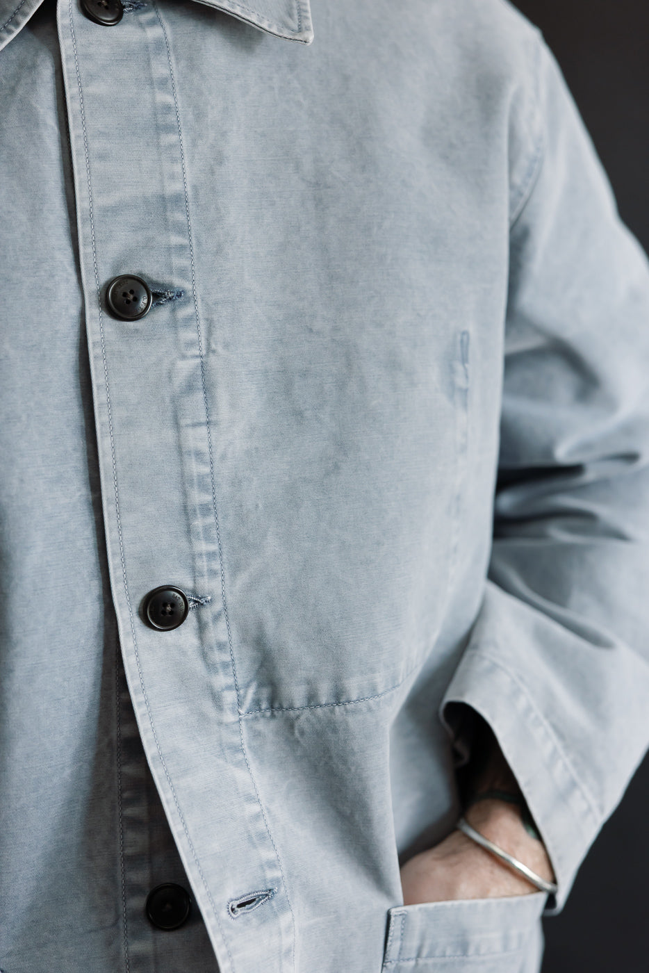 Close-up of someone wearing the M-B-006 Coach Jacket Nebraska Cotton in Coastal Blue, showcasing its luxurious hand-feel, three dark buttons, and relaxed fit with visible hip pocket stitching and a silver bangle.