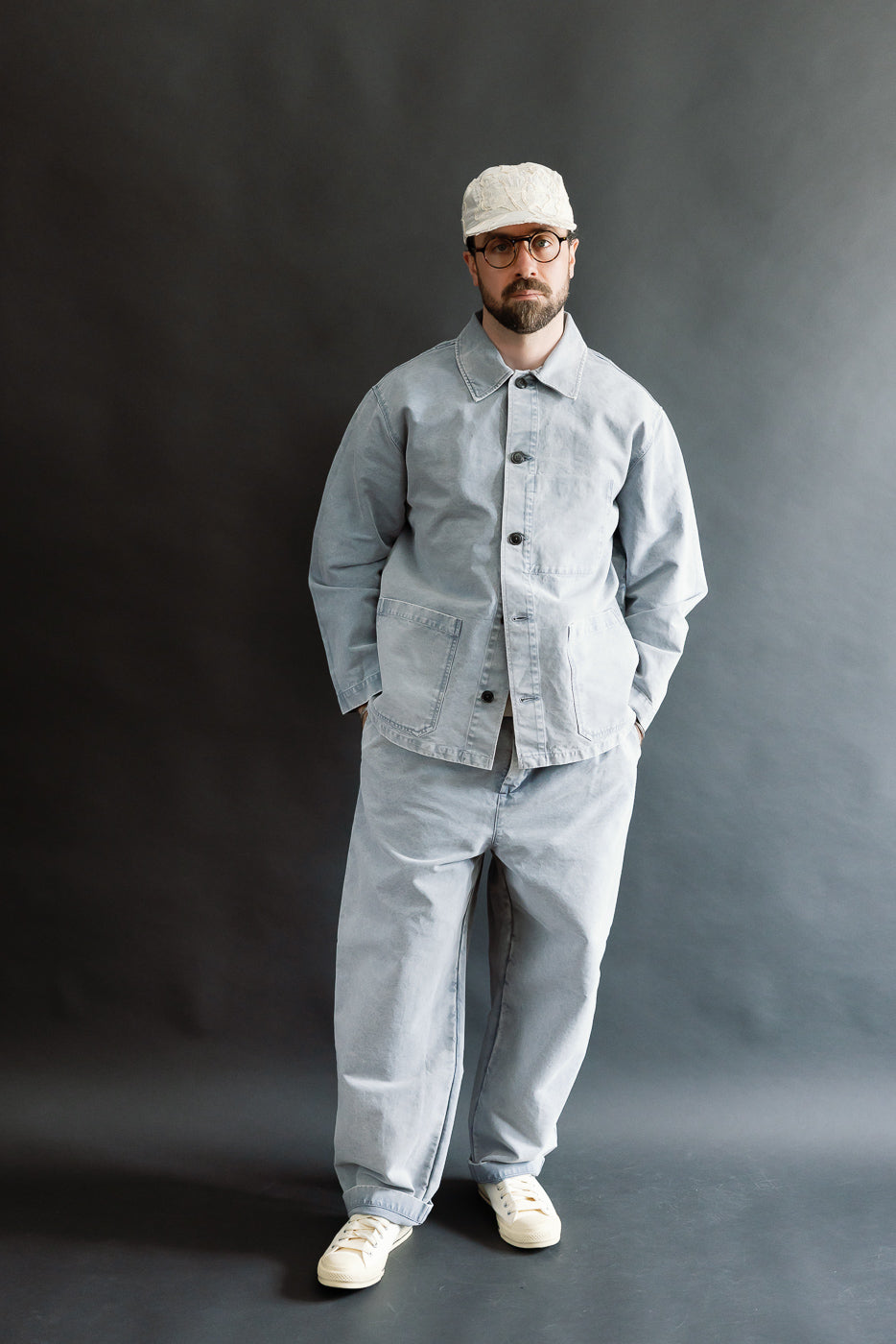 A 5'9 Caucasian male wearing the M-B-006 Coach Jacket Nebraska Cotton in Coastal Blue, styled with matching trousers, off-white canvas sneakers, a white cap, and tortoiseshell glasses, hands in jacket pockets.
