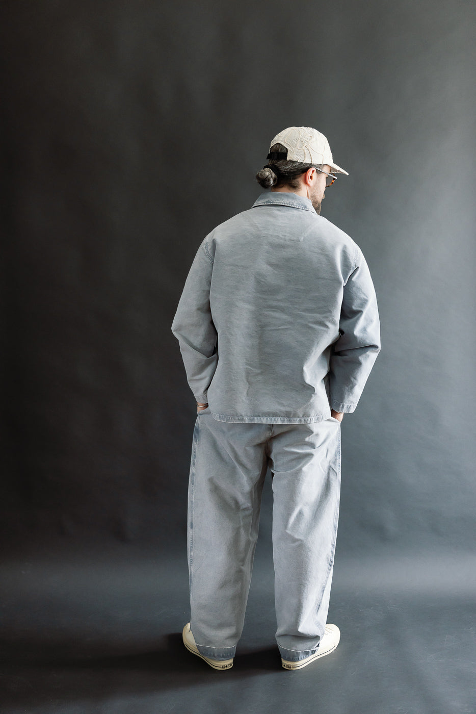 A person models the M-B-006 - Coach Jacket Nebraska Cotton - Coastal Blue, showcasing its relaxed fit and slightly dropped shoulders from the back view, paired with matching trousers and off-white sneakers.
