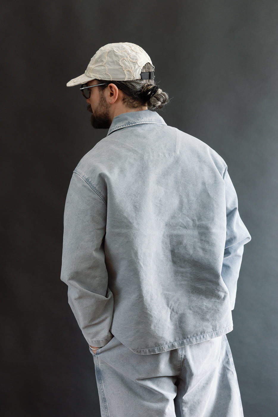 Person in a light grey M-B-006 Coach Jacket Nebraska Cotton - Coastal Blue, with relaxed fit and matching trousers, styled casually with a low bun, cap, and sunglasses against a plain backdrop.
