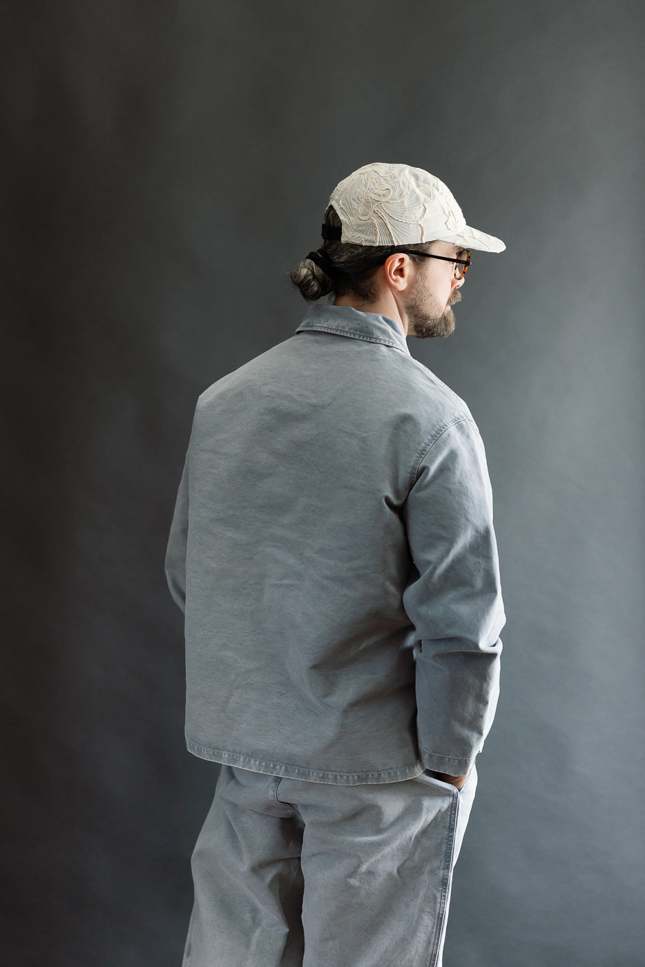 Man in profile wears M-B-006 Coach Jacket Nebraska Cotton - Coastal Blue, showing relaxed fit and slightly dropped shoulders, complemented by matching trousers, sunglasses, and a cream baseball cap.