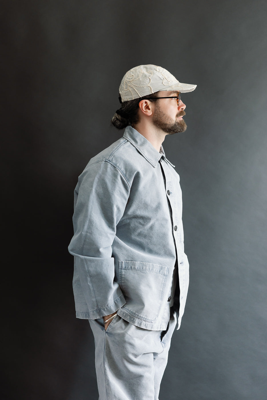 Man in profile wearing M-B-006 - Coach Jacket Nebraska Cotton - Coastal Blue, styled with relaxed fit and slightly dropped shoulders. He pairs it with matching trousers, embodying a modern workwear aesthetic.