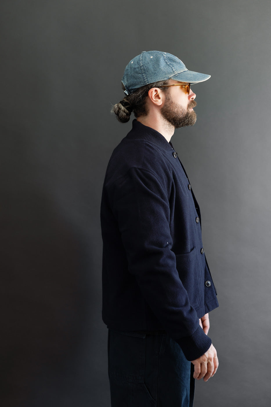 Side profile of a male model with an athletic build wearing a blue denim cap and navy bomber jacket.