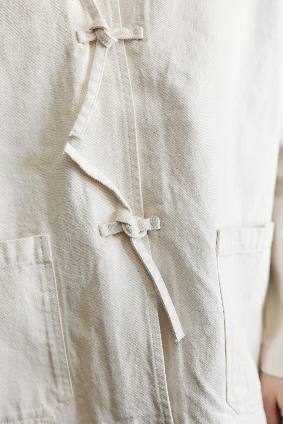 Close-up of front left side of ecru men's jacket, showing fabric texture, patch pockets, and knotted tie closure.