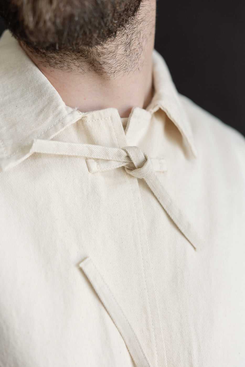 Close-up of front collar and tie closure detail on ecru jacket, worn by male model, 5'9
