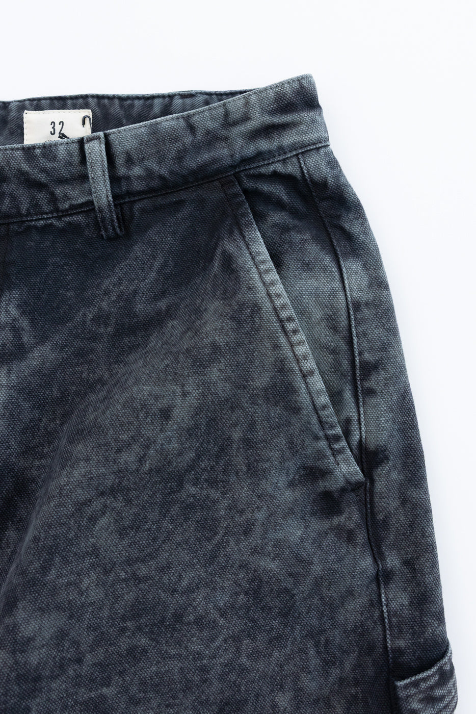 Close-up of front left side of asphalt canvas chino pants, showing waistband, belt loop, size tag, pocket, and textured fabric details.