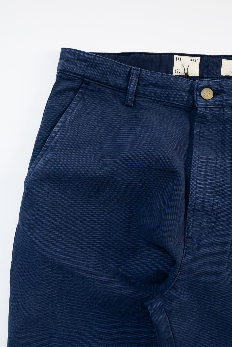 Close-up front view of Eat Dust Oakland chino indigo blue canvas pants, showing waistband, belt loops, front pocket, and metal button details, contemporary casual menswear at Dant.