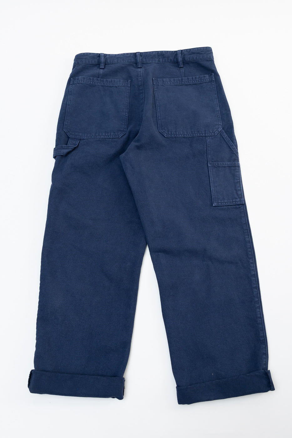 Flat lay of indigo blue canvas chino pants, back view, showing rear pockets and cuffed hems.