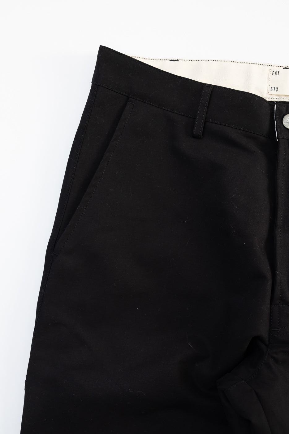 Close-up of front left side of black cotton chino pants, pocket and waistband details visible