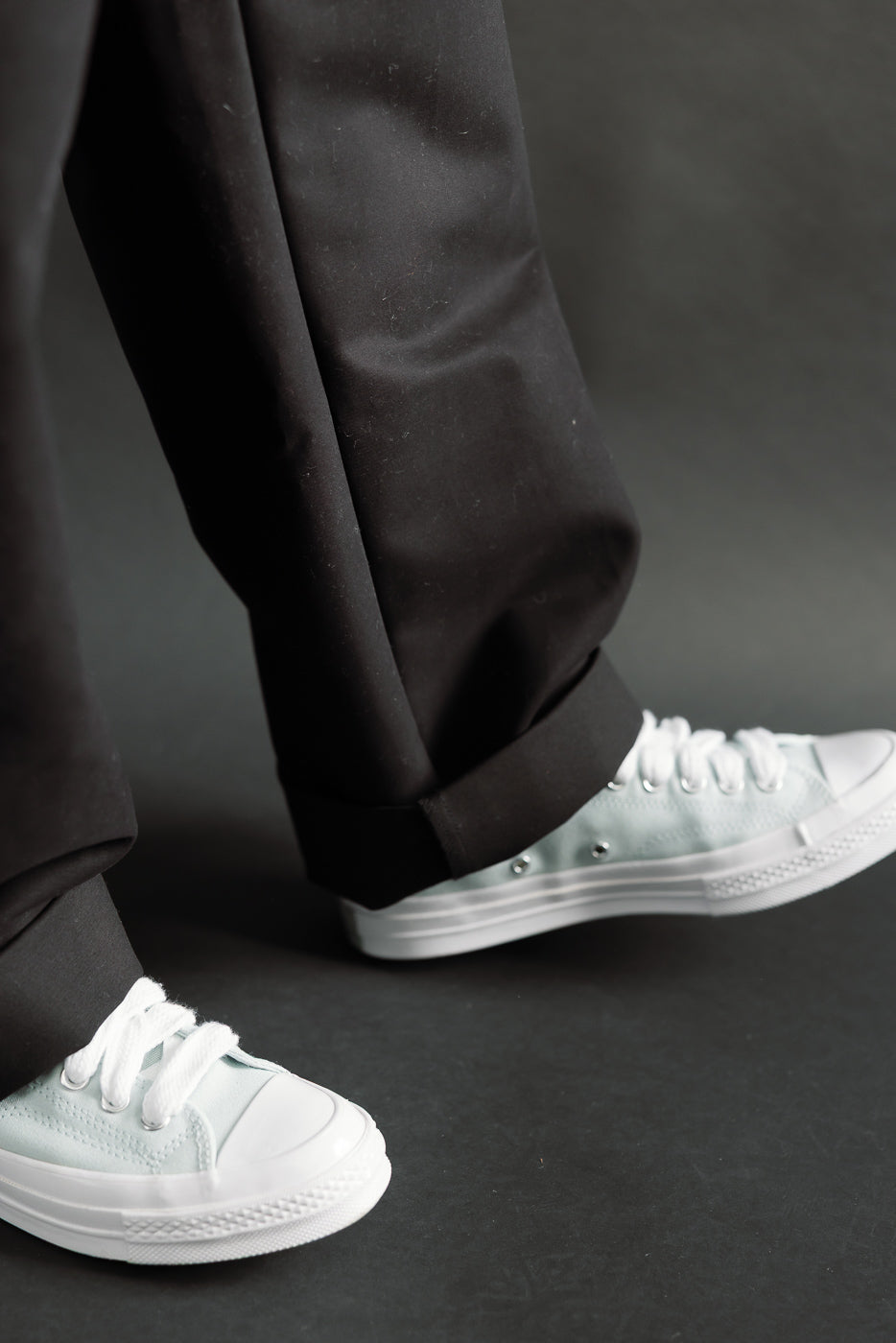 Close-up front view of male model's cuffed black cotton chinos and white sneakers. Model is 5'9