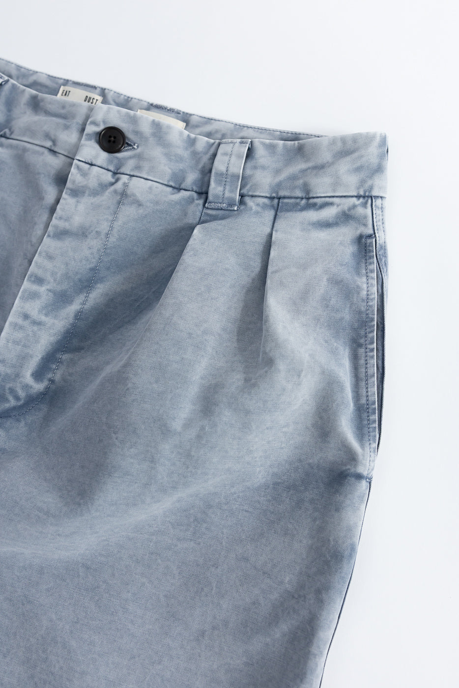 Close-up of front left side of light blue cotton chino pants, showing button, pleats, and belt loop details.
