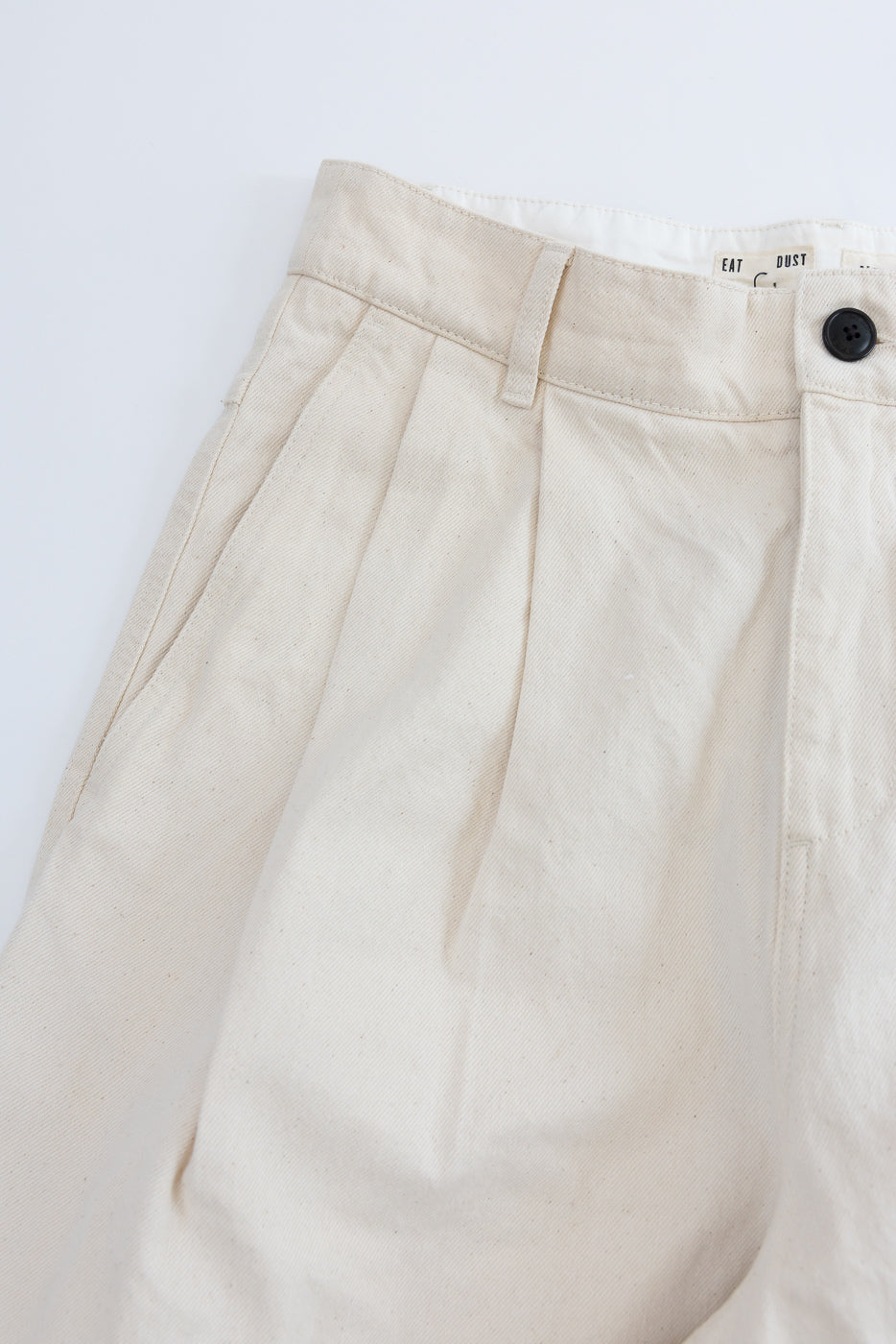 Close-up of front upper section of ecru canvas pleated trousers, showing waistband, belt loop, black button, side pocket, and visible tag details.
