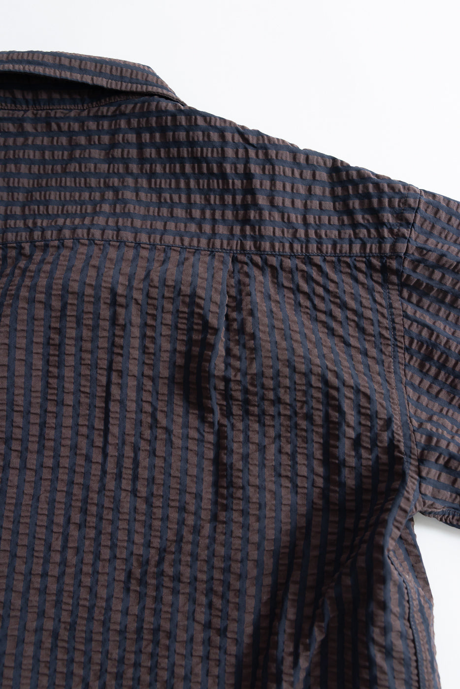 Close-up of the upper back and shoulder of a seersucker denim shirt in coffee and navy stripes, showing fabric texture and yoke detail.