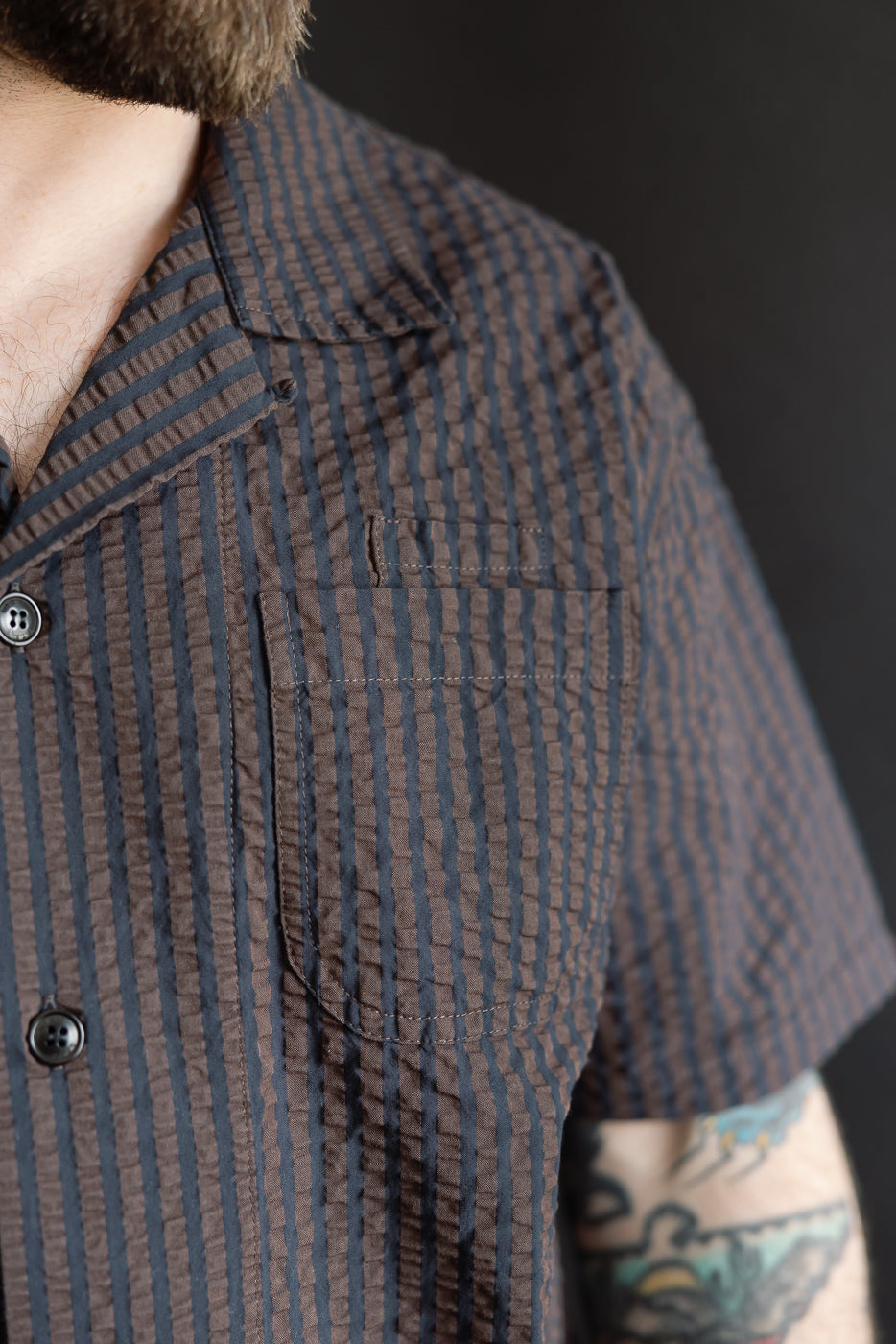 Close-up front view of male model wearing coffee navy seersucker Aloha shirt, showing chest pocket, textured fabric, notched collar, and short sleeves. Model is 5'9