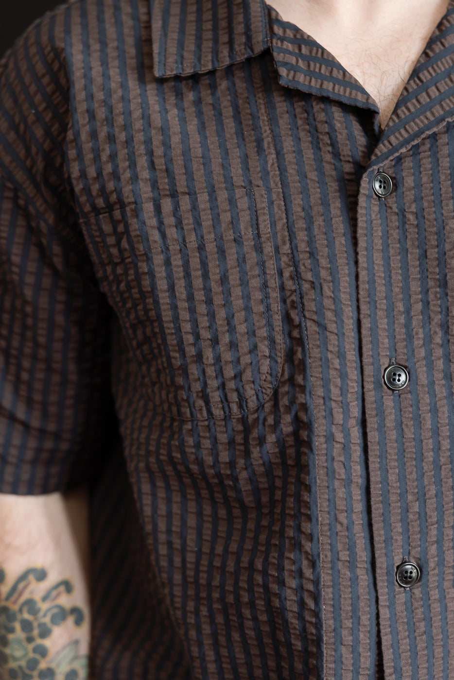 Close-up of front chest and collar of men’s coffee and navy striped seersucker denim short-sleeve aloha shirt on a male model, 5'9