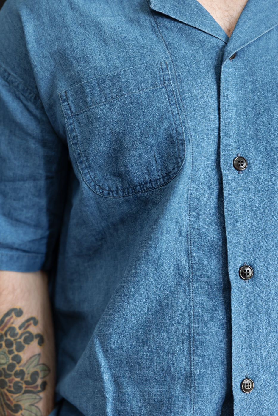 Close-up front of blue linen cotton denim aloha shirt on male model, showing chest pocket, buttons, and short sleeve.