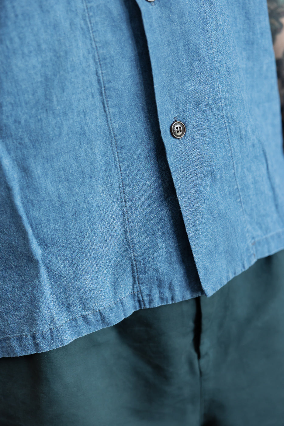Close-up of front lower section of blue linen-cotton denim aloha shirt showing button detail and fabric texture, worn by male model, 5'9