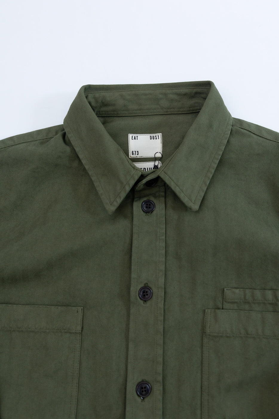 Close-up of the front collar and neckline of an olive green shirt showing the Eat Dust tag.