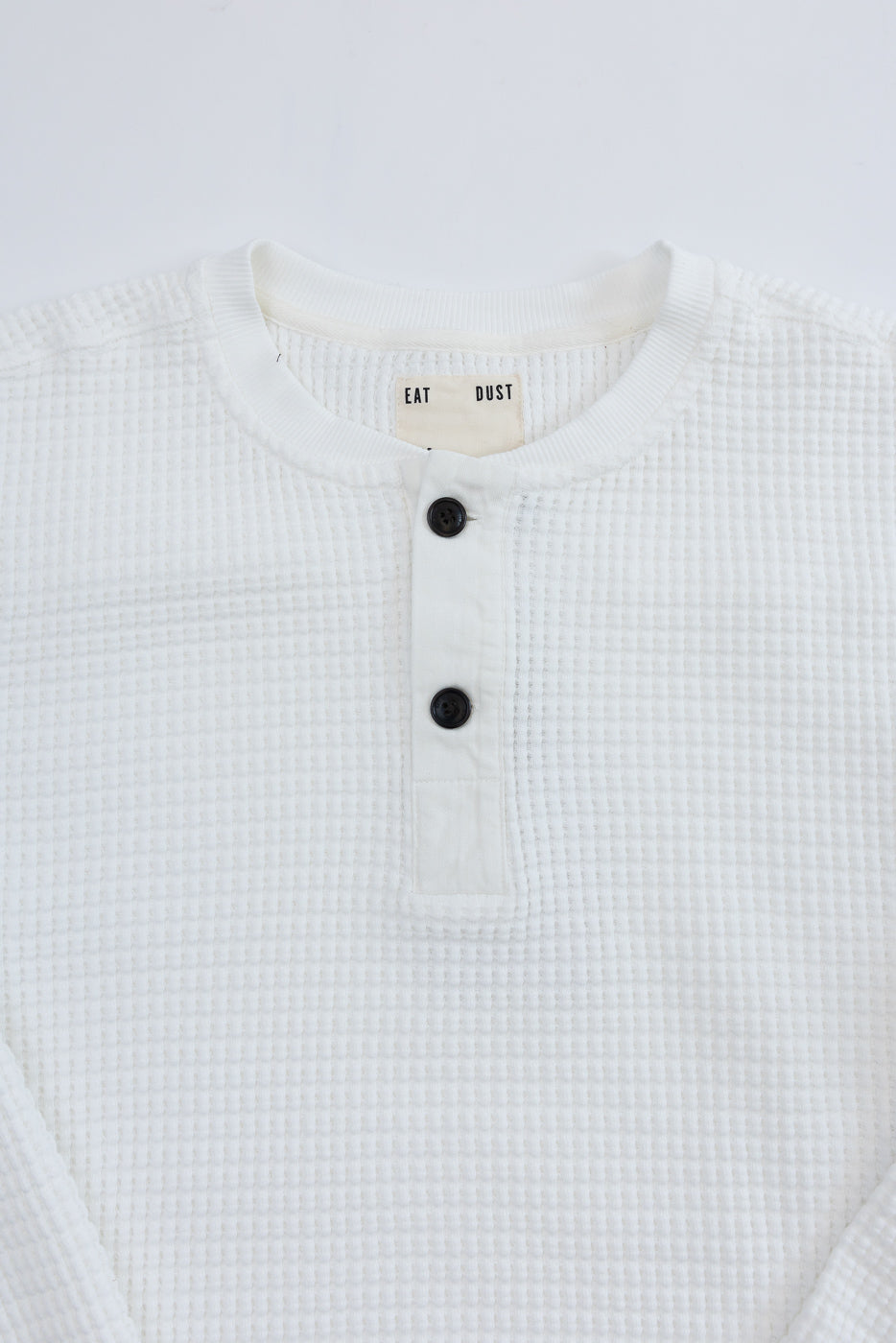 Close-up front view of white waffle knit henley with black buttons and Eat Dust tag.