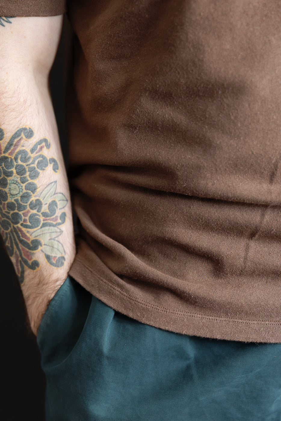 Close-up of male model’s left side, brown cotton hemp t-shirt hem and tattooed arm.