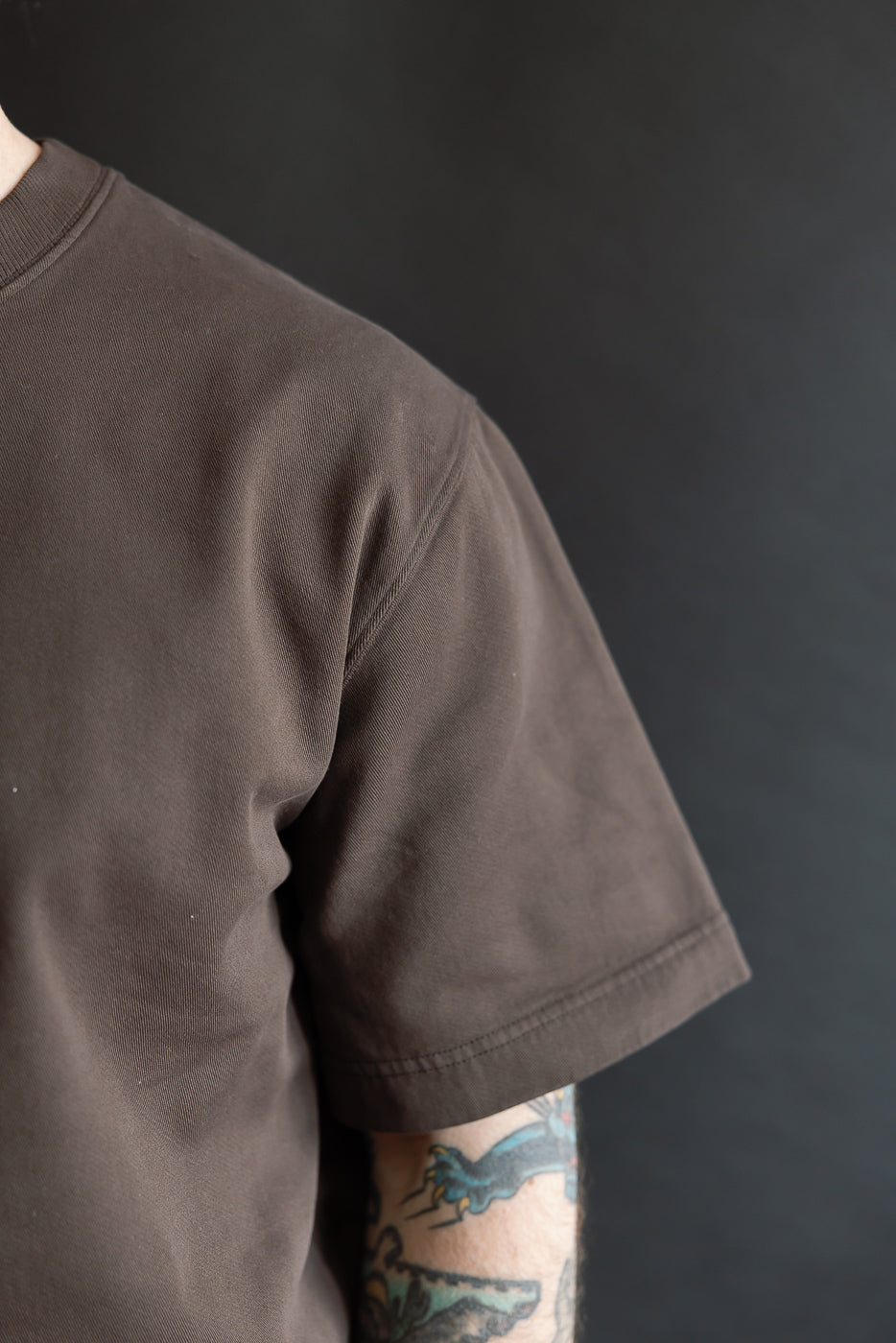 Close-up of male model, 5'9, 165lbs, athletic build, wearing coffee brown short sleeve t-shirt, left shoulder and sleeve detail