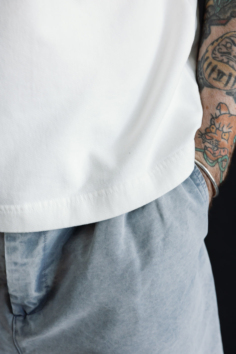 Person wearing M-J-009 - Power T Twill Jersey - Milk, a plain white T-shirt with a thick ribbed collar, showcasing a boxy cut and drop shoulder, highlighting colorful tattoos on their arm against a dark background.