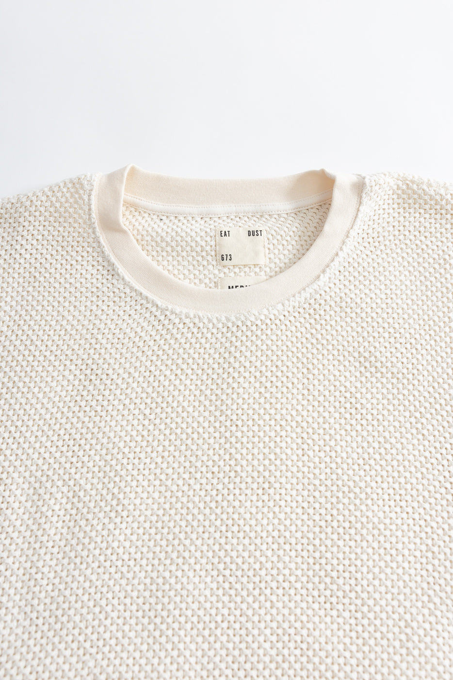 Close-up of front upper chest and collar on ecru textured cotton crewneck t-shirt, flat lay, showing ribbed neckline and woven fabric detail.