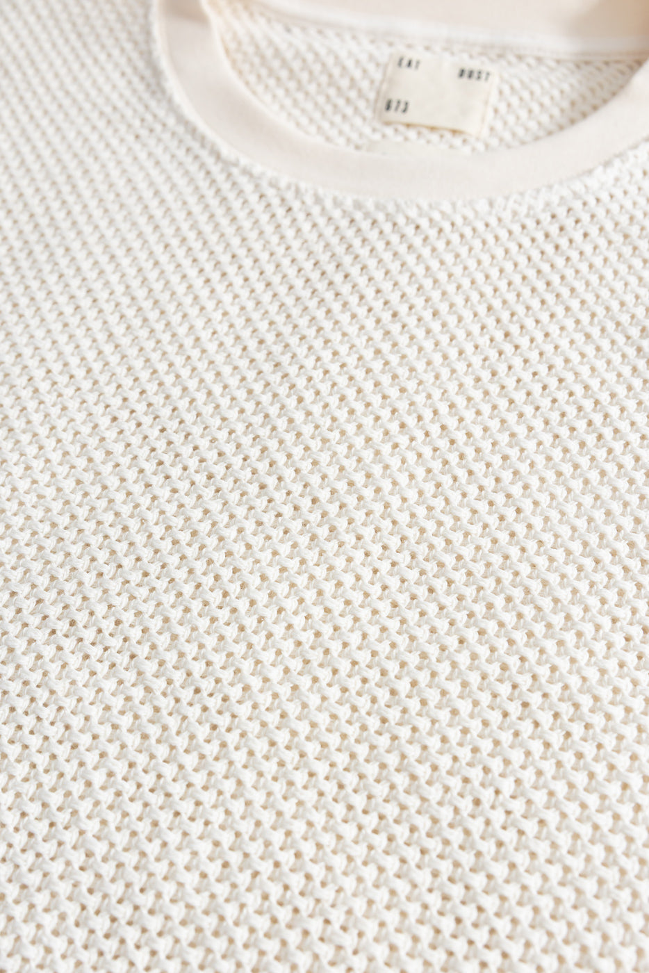Close-up of front chest detail, ecru textured cotton knit shirt, shows fabric weave and neckline tag.
