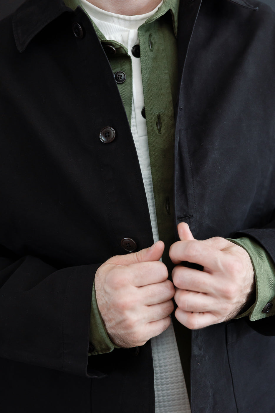 Close-up front view of a male model wearing a black trench coat over an olive green shirt.