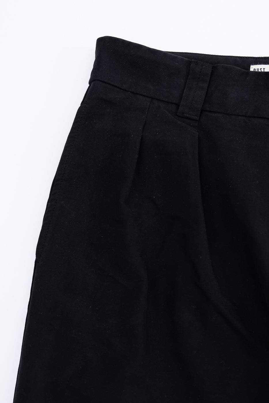 Close-up of the front waistband and pleated fabric of black trousers, showing the tag detail.