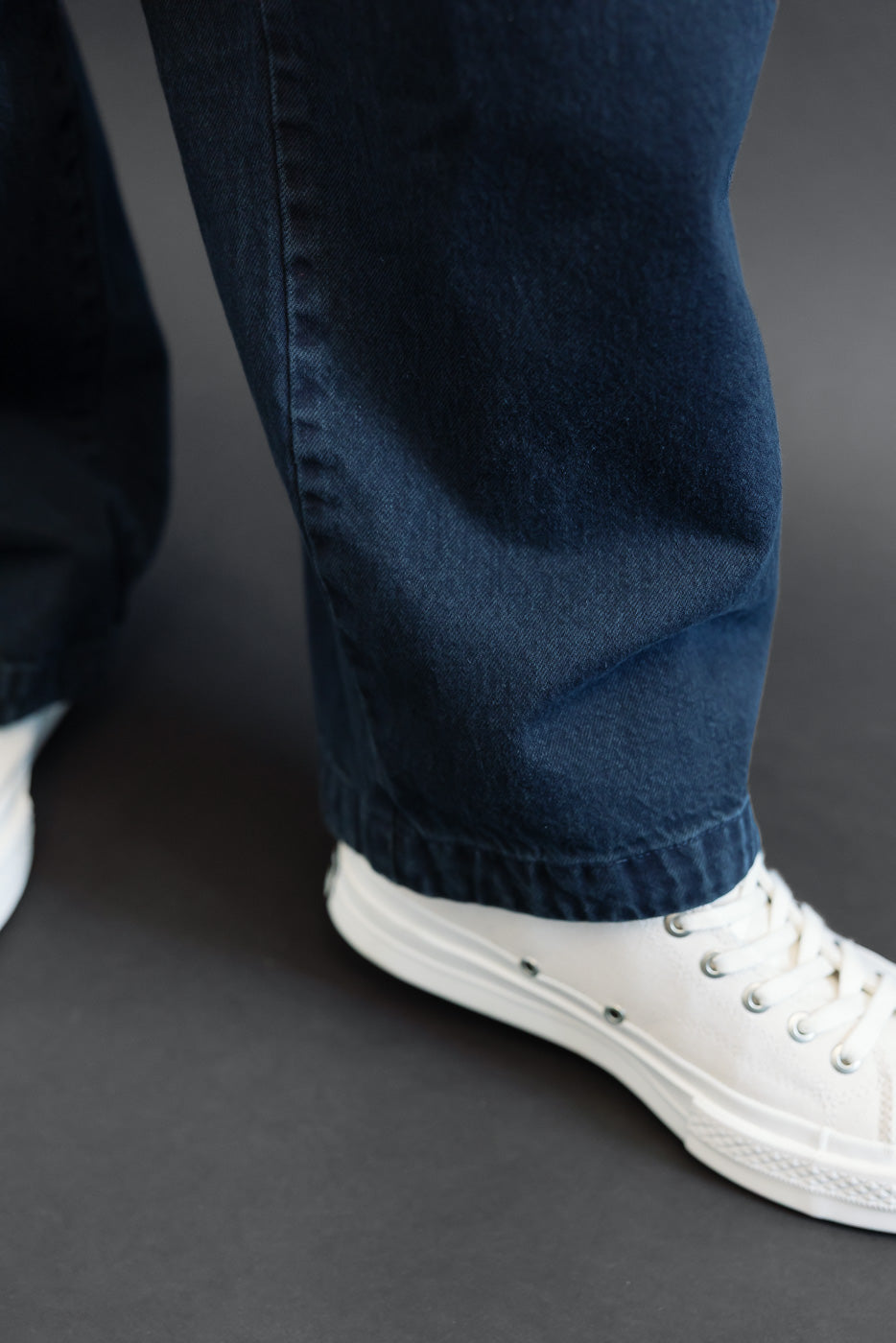 Close-up of the bottom hem and side of dark blue denim trousers worn by a male model with white sneakers.