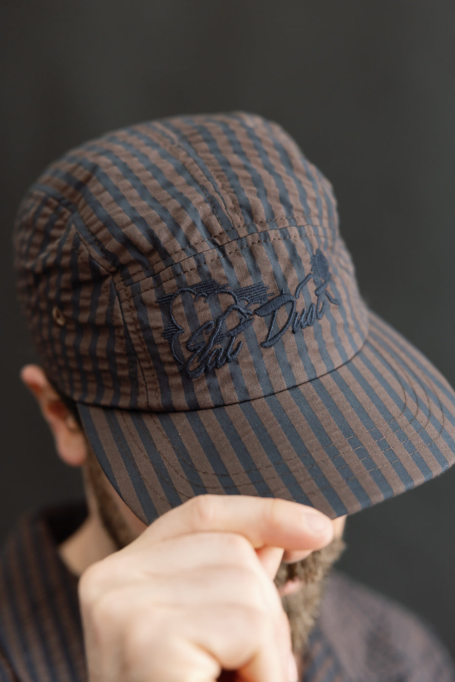 Close-up front view of a brown and navy striped 5-panel cap with embroidered logo, worn by a male model, 5'9