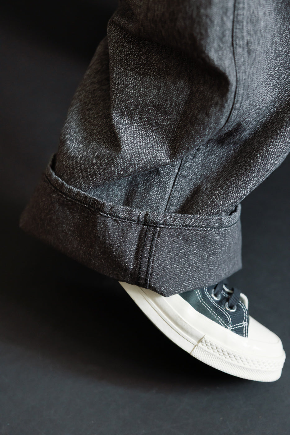Close-up side view of dark selvedge denim pant cuff, showing hem detail and texture, worn with dark sneaker.
