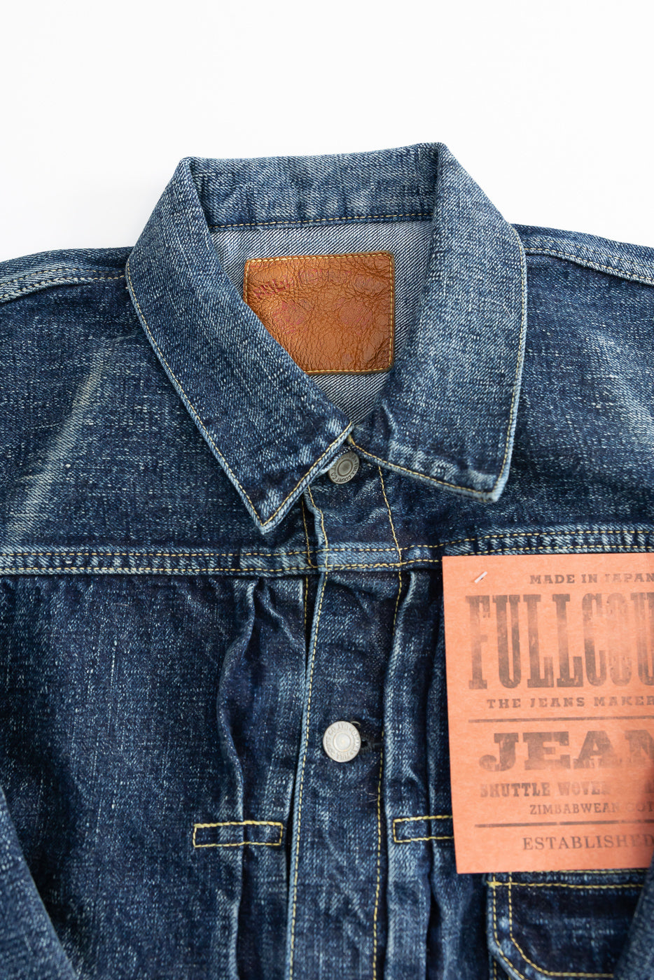 Close-up front view of hard washed indigo denim jacket, showing collar, button, yoke, and leather patch detail.
