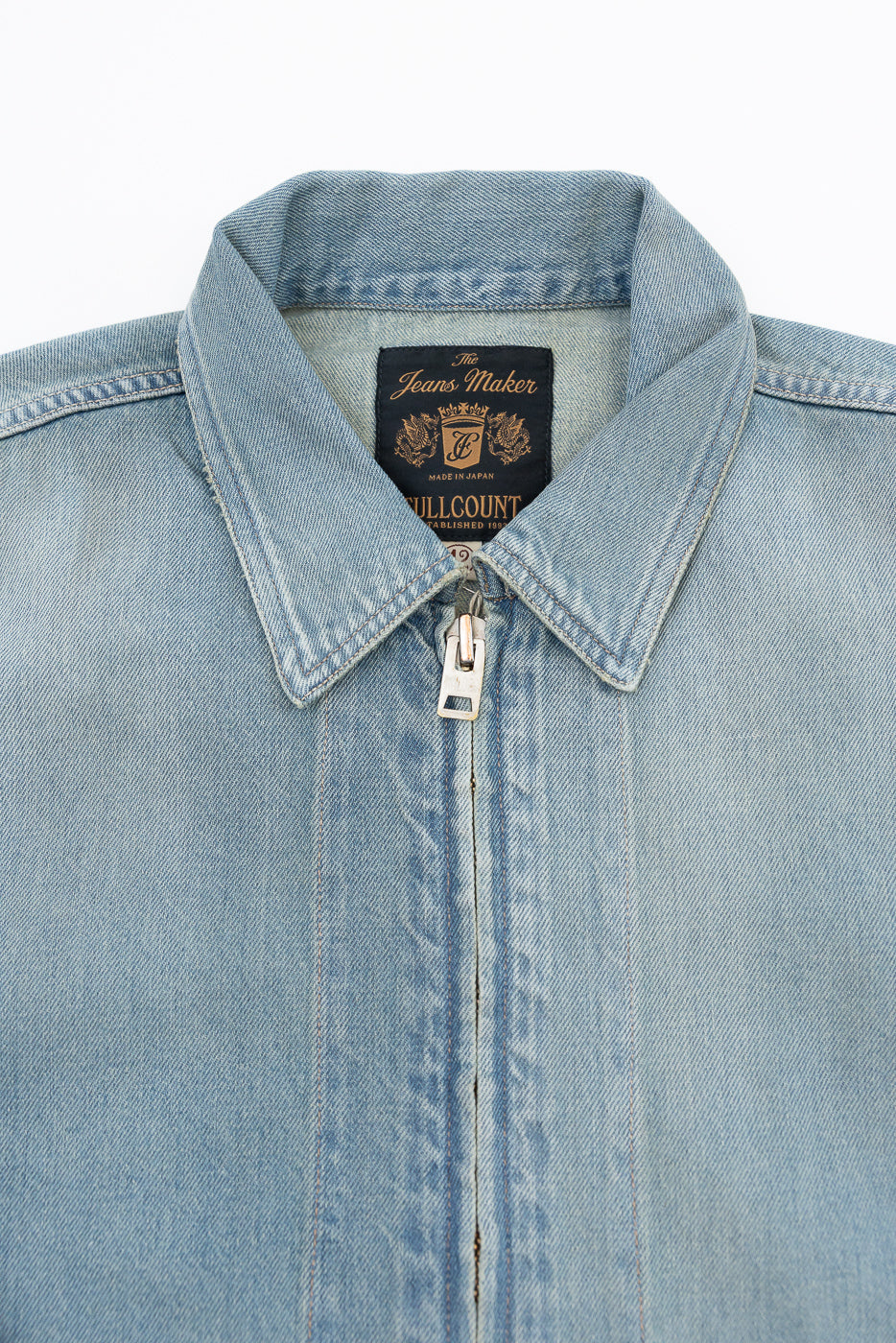 Close-up front view of light denim jacket collar, zipper, and 'The Jeans Maker' tag, showcasing fabric texture.