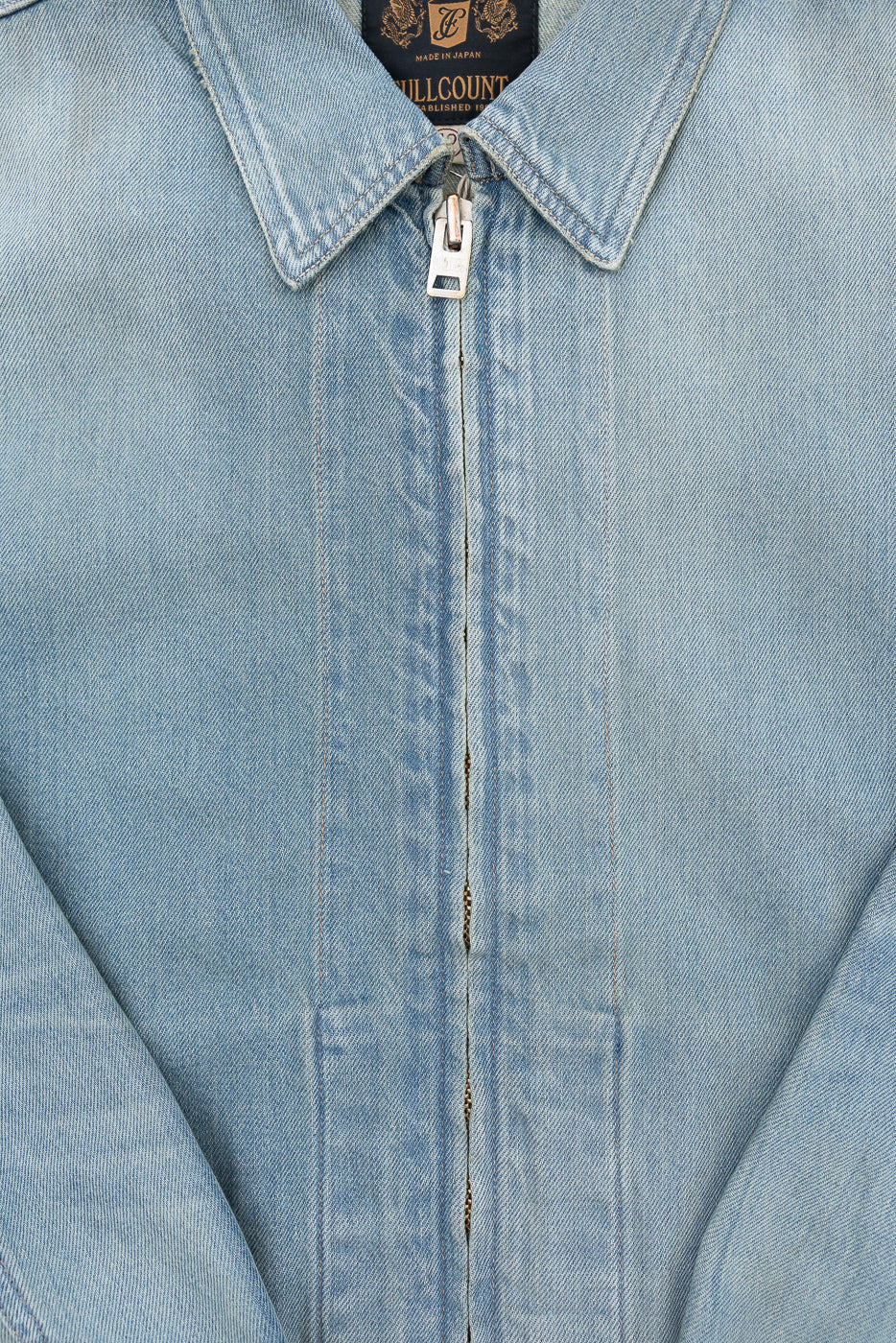 Close-up front view of light wash denim jacket with zipper, collar, and 'Fullcount' tag, showcasing fabric texture and stitching details.