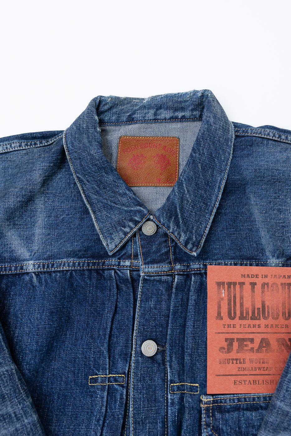 Heavily washed indigo denim jacket with a visible full count label on the left chest pocket on a white background, close up of collar and leather nape tqag with full count red logo. 