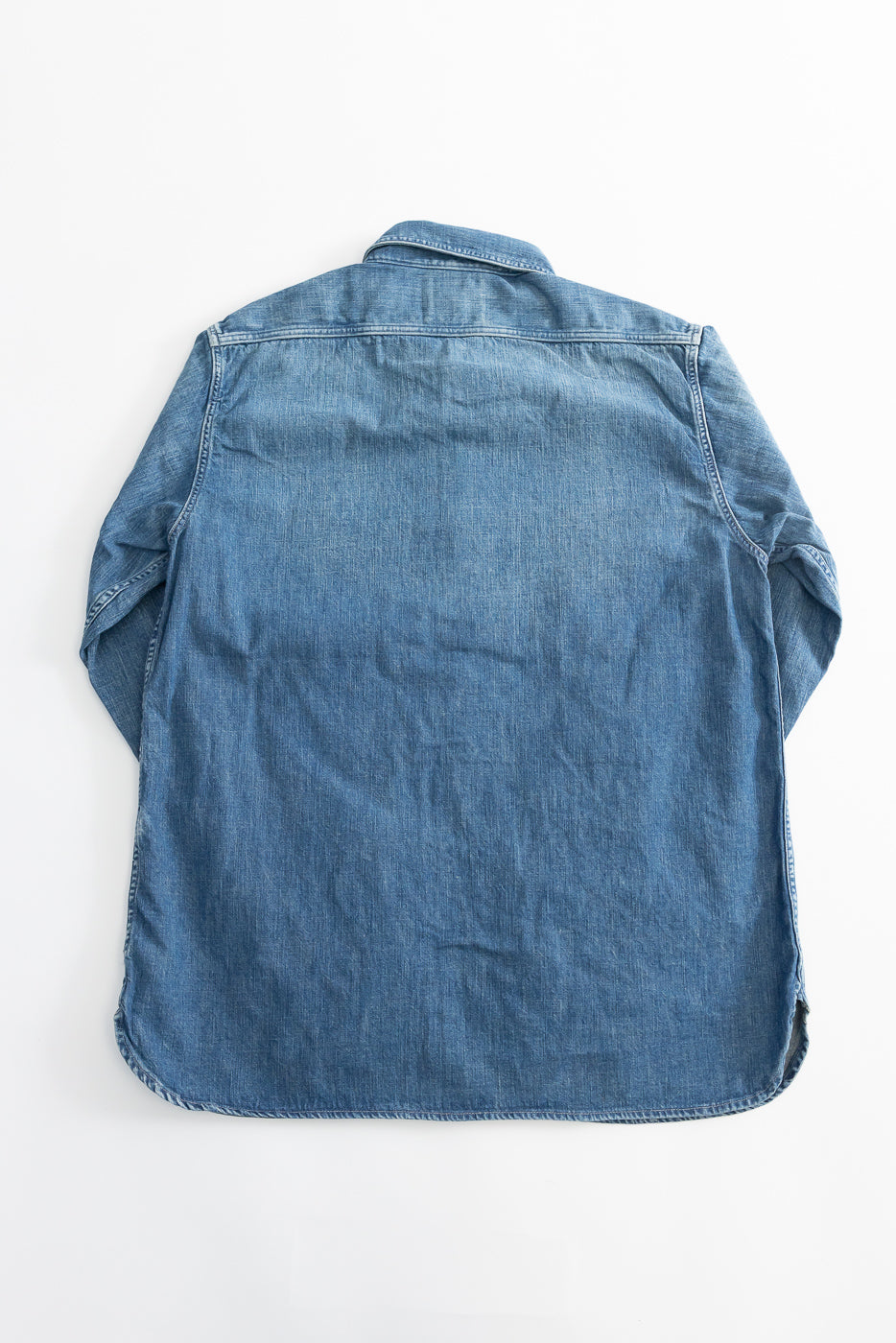 Flat lay of back side of indigo denim work shirt, showing full garment with curved hem.