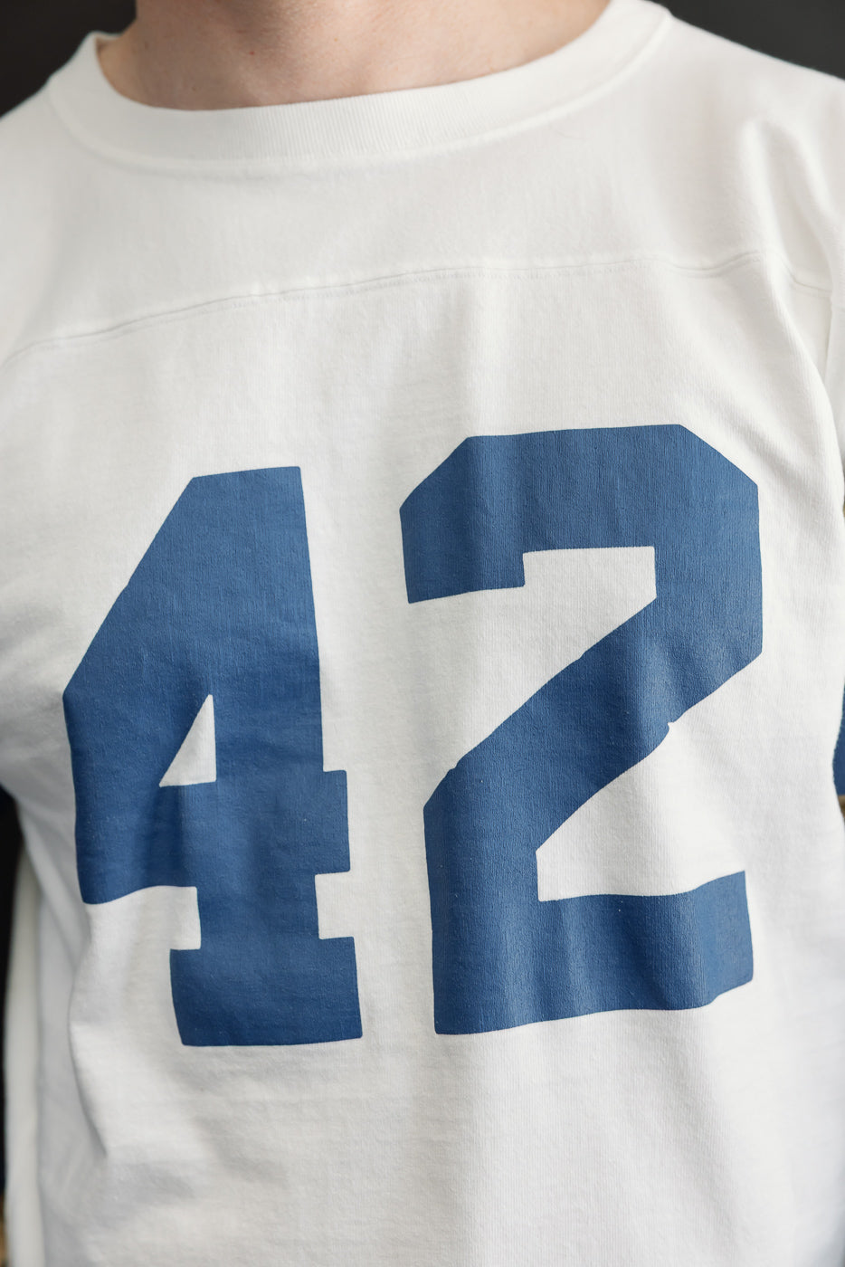 Close-up front view of white crewneck tee with bold blue '42' print, worn by male model, 5'9