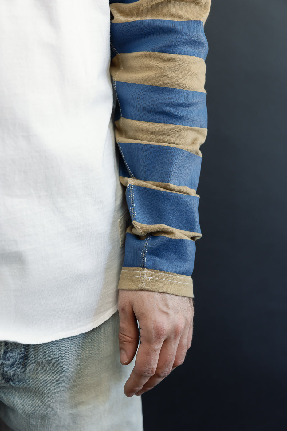 Close-up side view of male model's arm in striped long sleeve shirt, showing blue and tan ribbed fabric, contrast stitching, and cuffed sleeve detail.