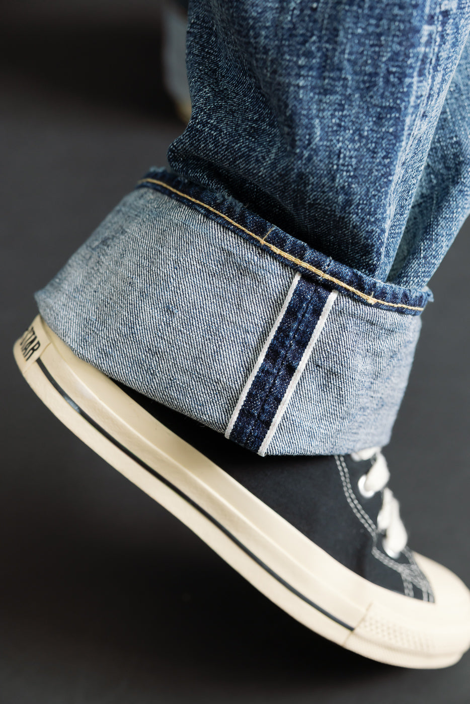 Close-up of male model's lower leg wearing hard washed indigo selvedge denim jeans, cuff detail and sneaker, showing leg side view. Model is 5'9